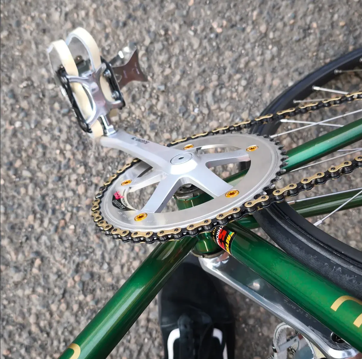 Detail der Sugino-Kurbel in Silber mit vergoldeten Kettenblatt-Schrauben und goldener Fahrradkette, Blick von oben auf Tretlager und Pedal.