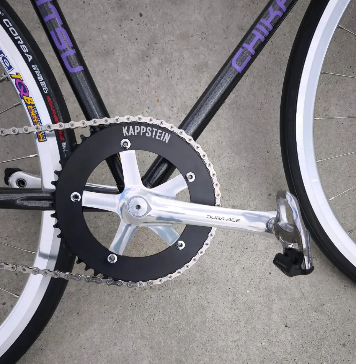 Nahaufnahme der Dura-Ace-Kurbel mit schwarzem Kappstein-Kettenblatt und silberner Fahrradkette, Vittoria-Corsa-Reifenaufschrift im Hintergrund.