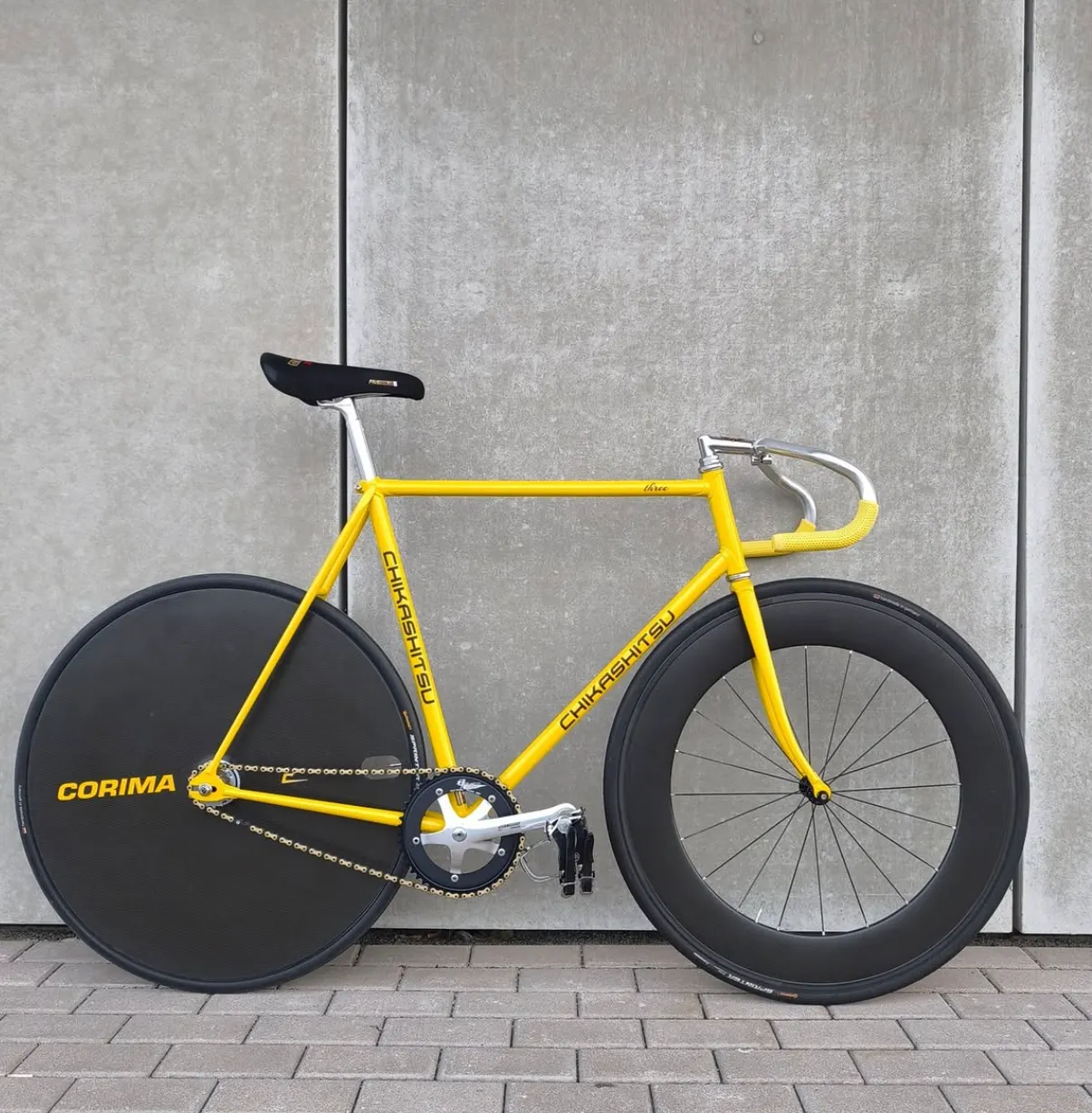 Gelber Bahnrad-Rahmen Chikashitsu Three von der Seite vor Betonwand, Corima-Scheibenrad hinten, Rennlenker mit gelbem Lenkerband.