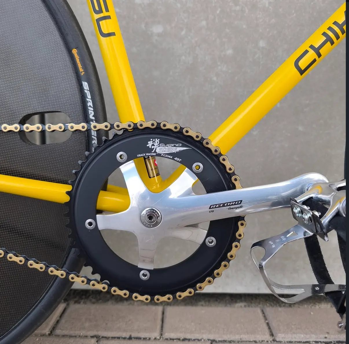 Nahaufnahme der Campagnolo-Record-Kurbel mit schwarzem Sugino-Zen-Kettenblatt und goldener Kette vor dem Corima-Scheibenrad.