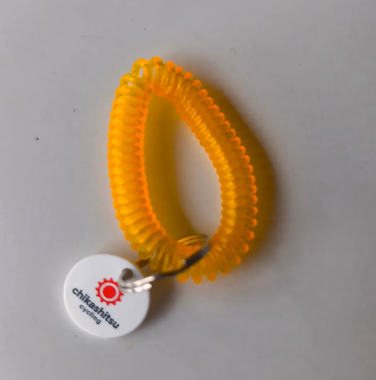 Gelbes Spiral-Schlüsselband mit rundem weißem Anhänger, darauf das rote Chikashitsu-Cycling-Kettenblatt-Logo.
