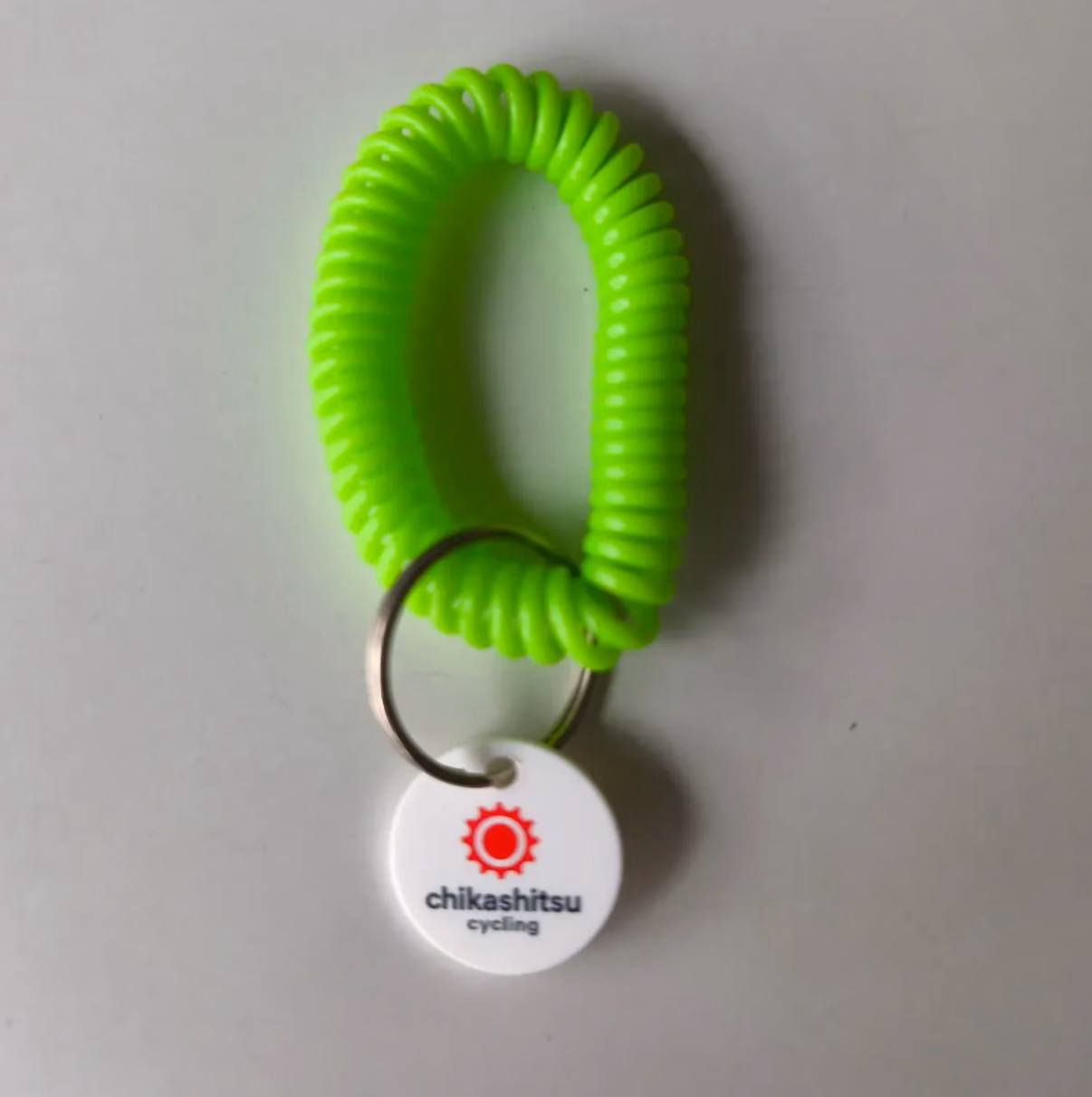 Neongrünes Spiral-Schlüsselband mit Schlüsselring und rundem Chikashitsu-Cycling-Logo-Anhänger auf hellem Untergrund.