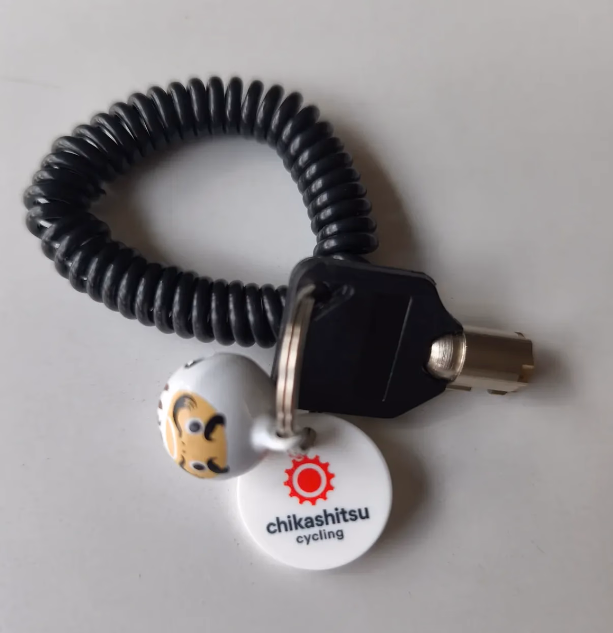 Schwarzes Spiral-Schlüsselband mit Fahrradschlüssel, kleiner Daruma-Figur und rundem Chikashitsu-Cycling-Logo-Anhänger.