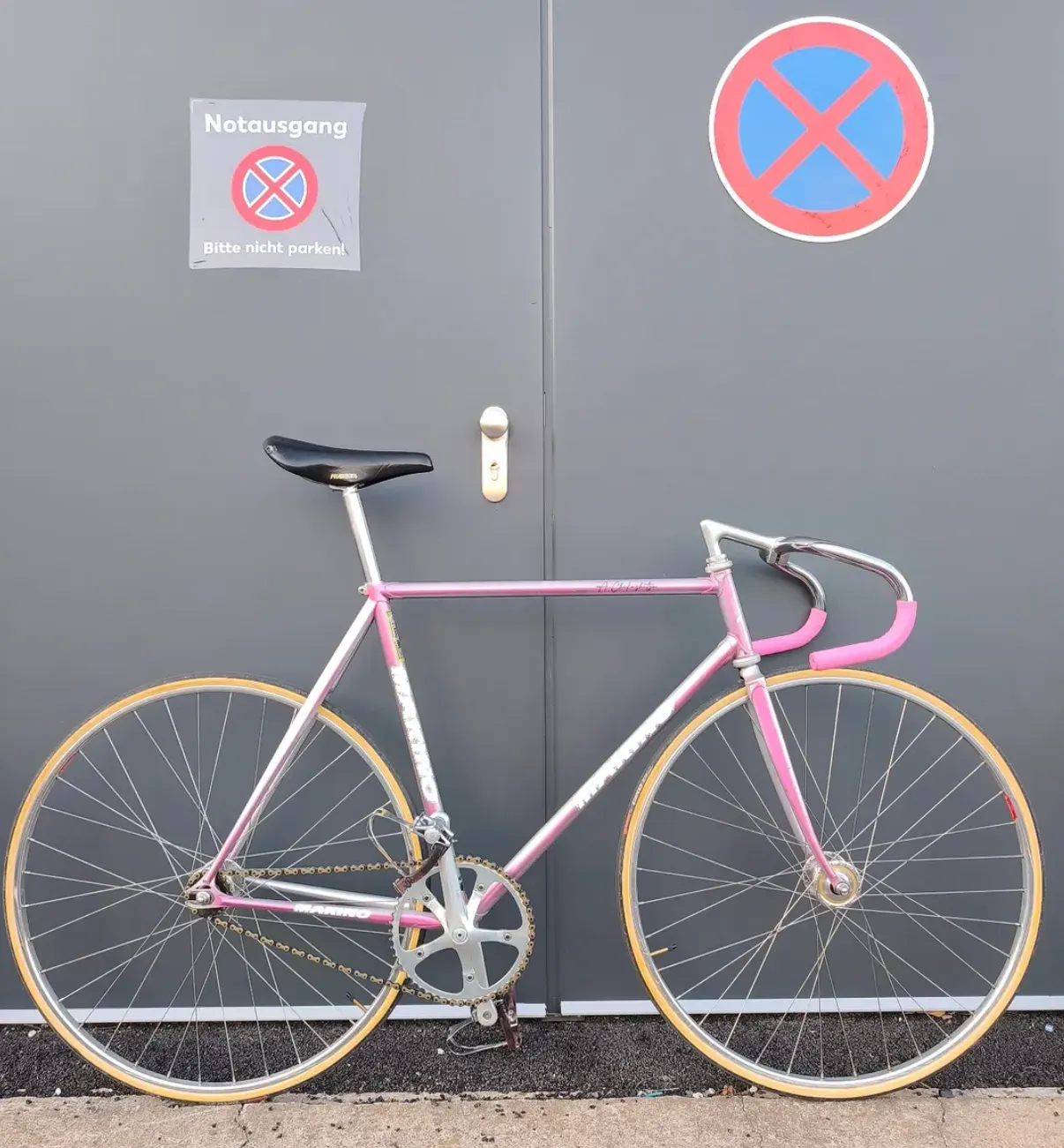 Pink-silbern schimmerndes Marino-Rennrad vor einer grauen Toreinfahrt mit Notausgang-Schild, schwarzer Sattel, gelbe Reifen.