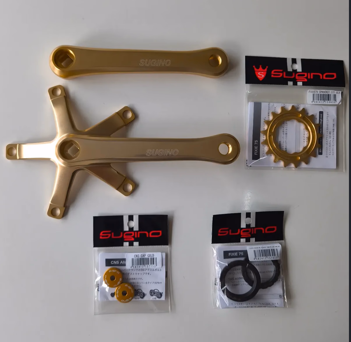 Gold eloxierte Sugino-Kurbelarme und ein Sugino-Fixie75-Ritzel, original verpackt, auf weißem Untergrund arrangiert.