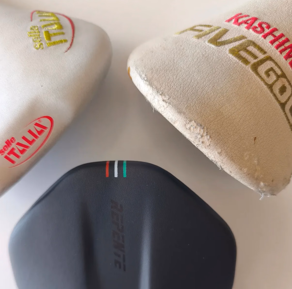 Drei Fahrradsättel von oben: cremefarbener Selle-Italia-Flite-Sattel, cremefarbener Kashimax-Avegol-Sattel und ein schwarzer Sattel mit Trikolore-Streifen.